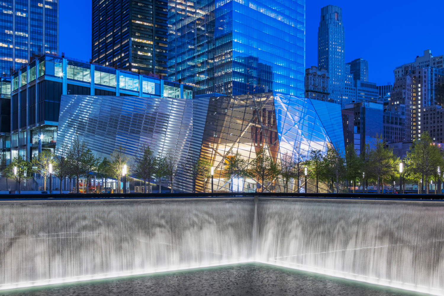 Nighttime photograph of an architecturally dramatic glass building with sunken reflecting pool in the foreground. 